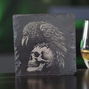 May include: A dark grey slate coaster featuring an engraved design of an eagle and a skull. A glass of amber liquid is to the right. The coaster is a decorative item for a bar or home, with a gothic theme.