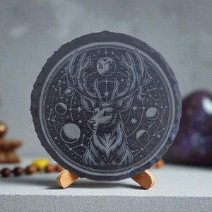 Pode incluir: Uma base para copos redonda de ardósia cinza escuro com uma gravura detalhada de um veado com grandes chifres, rodeado por elementos celestes. A base para copos é exibida em um pequeno suporte de madeira, com um fundo desfocado, incluindo uma estátua dourada de Buda e pedras decorativas.