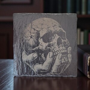 May include: A square, grey slate coaster with a white illustration of a human skull held in a hand. A lit candle in a brass holder sits on a book next to the coaster. The background includes a bookshelf.