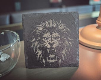 Puede incluir: Posavasos cuadrado de pizarra gris oscuro con una imagen grabada de la cara de un león rugiendo. La melena y los rasgos faciales del león están representados en un gris más claro. El posavasos está sobre una superficie de madera.