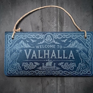 May include: A rectangular dark grey slate sign with a jute rope hanger. The sign features an engraved design with the text "WELCOME TO VALHALLA," flanked by two ravens, a Viking ship, mountain silhouettes, and a Valknut symbol at the top.