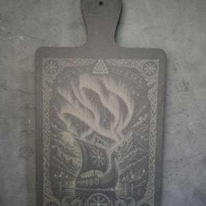 May include: A dark grey rectangular wooden serving board with a handle and a hole for hanging. The surface features a laser-engraved illustration of a Viking longship on water, mountains, and aurora borealis, framed by intricate knotwork and runes.