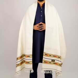 May include: Cream-colored shawl with a decorative border featuring red, green, and gold accents. The shawl has a fringed edge and is draped over a dark blue garment. The border includes a small rectangular label.