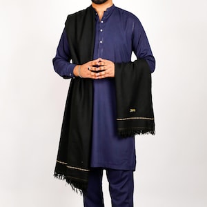 May include: A black shawl with a fringed edge is draped over a navy blue outfit. The shawl has a gold-colored border and a small tag with text. The outfit consists of a long-sleeved shirt and matching pants.