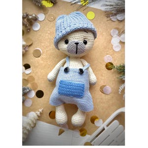 Może przedstawiać: Szydełkowy miś w jasnoniebieskiej czapce i ogrodniczkach. Miś ma kremowy korpus, szary pyszczek i czarne guziczkowe oczy. Ogrodniczki mają przednią kieszeń. Tekst "Wzór „Miś Polarny"" znajduje się na dole.