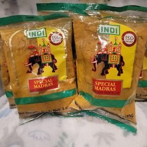 Puede incluir: Se muestran varios paquetes de polvo de curry Special Madras. Cada paquete presenta un diseño amarillo y verde con una ilustración de un elefante que lleva una estructura decorativa. El texto "INDI" y "SPECIAL MADRAS" son claramente visibles.