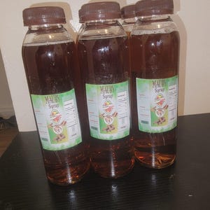 May include: Several clear plastic bottles filled with a dark brown liquid, each capped with a brown lid. The bottles have green and white labels that read "MAUBY Syrup". The bottles are arranged on a dark surface.