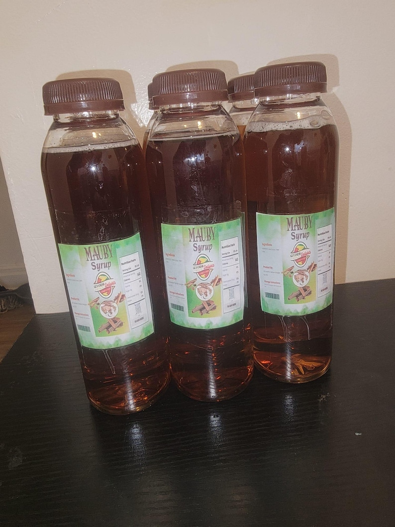 May include: Several clear plastic bottles filled with a dark brown liquid, each capped with a brown lid. The bottles have green and white labels that read "MAUBY Syrup". The bottles are arranged on a dark surface.