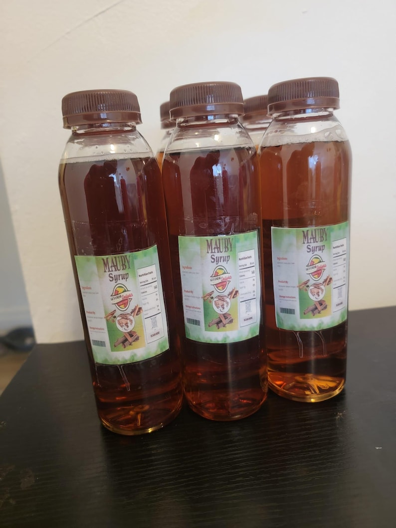 May include: Four clear plastic bottles filled with a brown liquid, each topped with a brown cap. The bottles have labels with the text "MAUBY Syrup" and a graphic. The bottles are arranged on a dark surface.