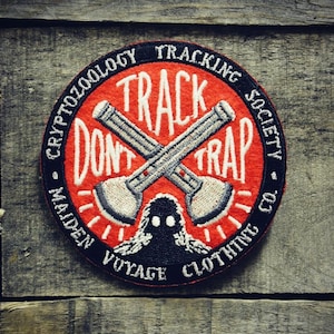 Op de afbeelding: Rode geborduurde patch met de tekst "TRACK DON'T TRAP" en een gekruist paar bijlen. De patch heeft ook de tekst "CRYPTOZOOLOGY TRACKING SOCIETY - MAIDEN VOYAGE CLOTHING CO."