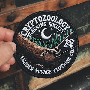 Op de afbeelding: Zwarte geborduurde patch met de tekst "Cryptozoology Tracking Society Maiden Voyage Clothing Co." en een halve maan met sterren. De patch toont een bruine hand die uit een bos scène reikt.