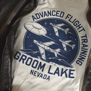 Pode incluir: Uma camiseta cinza claro com um design gráfico azul e branco que apresenta um OVNI e três aviões voando sobre uma cordilheira. O gráfico também inclui o texto "Advanced Flight Training", "Groom Lake" e "Nevada".