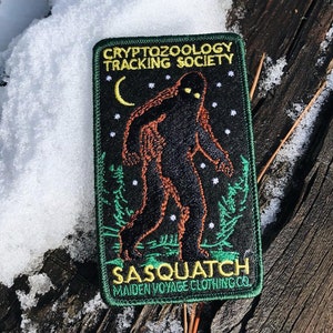Può includere: Toppa ricamata verde, nera e gialla con un'illustrazione di sasquatch, una luna crescente e stelle. Il testo recita "Cryptozoology Tracking Society" e "Sasquatch Maiden Voyage Clothing Co."