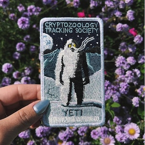 Puede incluir: Parche bordado con una ilustración de un yeti contra un fondo de montañas nevadas bajo un cielo estrellado. El parche dice "Cryptozoology Tracking Society" y "Yeti", con "Maiden Voyage Clothing Co." debajo.