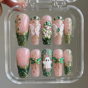 May include: A set of ten press-on nails in a clear plastic case, featuring a light pink base with intricate gold and dark green scrollwork, white 3D butterflies, green mushrooms, a white ghost, green dragonflies, and small green rhinestones.