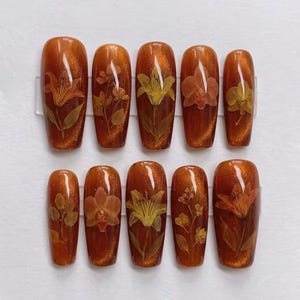 May include: A set of ten press-on nails featuring a glossy, deep amber cat-eye finish. Each nail is decorated with embedded, dried botanical elements, including various yellow and orange lily and orchid flowers, arranged in unique floral patterns.