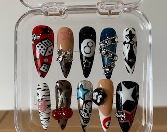 Op de afbeelding: Een set van tien lange, puntige valse nagels in een doorzichtige plastic doos. De nagels hebben verschillende ontwerpen, waaronder dobbelstenen, strikken, een achtbal, sterren en een schedel. Kleuren zijn rood, zwart, wit en nude.