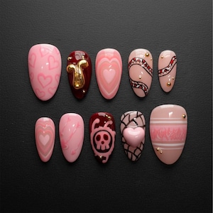 May include: A set of ten artificial nails in various designs. The nails feature pink and nude tones with heart, arrow, skull, and abstract patterns. Some nails have gold accents, and a 3D heart is present on one nail.