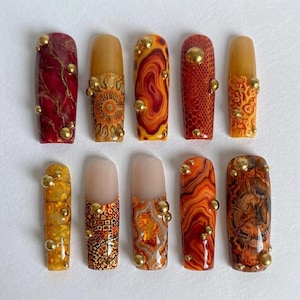 May include: A collection of ten artificial nails with various designs. The nails feature a range of colors, including red, orange, and yellow, with gold embellishments. Some nails have marble, reptile, or floral patterns, while others have a solid color.