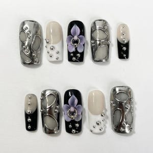 May include: A set of ten press-on nails featuring a variety of designs including metallic silver chrome finishes with raised grey accents, black and white bases with silver studs, and a central black nail adorned with a 3D purple flower charm.
