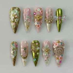 May include: A collection of ten long, pointed artificial nails. Each nail features intricate designs, including floral patterns, a bee, a dragonfly, and hummingbirds. Colors include pink, green, gold, and white, with detailed embellishments.