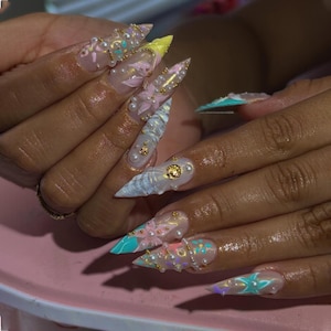 May include: Long stiletto-shaped artificial nails featuring a clear base with intricate 3D designs including pastel pink and blue floral patterns, iridescent accents, small white pearls, and tiny gold metallic studs applied across the surface.
