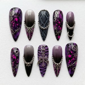 May include: A set of ten press-on nails featuring a pointed shape with purple and black color schemes. Designs include silver gear charms, barbed wire accents, snake skin patterns, ombre gradients, metal studs, and a dangling cross charm with crystals.