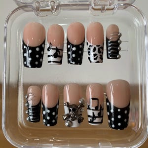 May include: A set of press-on nails in a clear plastic case. The nails feature a nude base with various designs, including black polka dots, musical notes, and a skull with rhinestones. Some nails have silver metal accents.