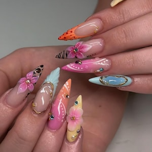 May include: A set of long, pointed acrylic nails with various designs. The nails feature a range of colors, including orange, pink, blue, and yellow, with floral and abstract patterns. Some nails have 3D flower embellishments and rhinestones.