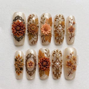 May include: A set of ten press-on nails with a shimmering cream base, featuring 3D relief designs of orange and brown autumn flowers, gold filigree frames, intricate gold scrollwork, maple leaves, and small white pearls arranged in various patterns.