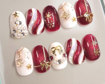 Könnte beinhalten: Ein Set aus zehn künstlichen Nägeln mit festlichem Design. Die Nägel weisen eine Kombination aus weißen, roten und goldenen Farben auf, mit Verzierungen wie Perlen, Schleifen und Schneeflockenmustern. Die Nägel sind oval geformt und haben ein glänzendes Finish.