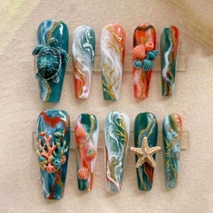 May include: A set of ten long, coffin-shaped artificial nails with ocean-themed designs. Each nail features a unique combination of teal, white, orange, and gold accents, with 3D elements like a turtle, coral, starfish, and seashells.