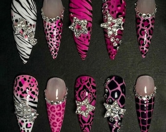Op de afbeelding: Een set van tien lange, puntige kunstnagels. Ontwerpen omvatten zebra strepen, luipaardprint en abstracte patronen in roze, zwart en wit. Sommige nagels hebben strass versieringen, waaronder sterren en randen.