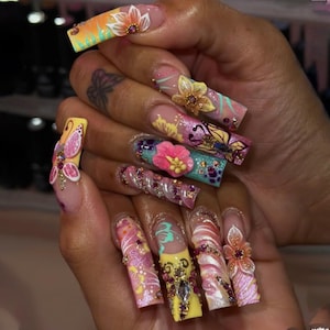 May include: A hand displaying long, decorated acrylic nails. Each nail features a unique design, including floral patterns, butterflies, and sparkling embellishments in shades of pink, yellow, orange, and blue. The nails are adorned with rhinestones and 3D elements.