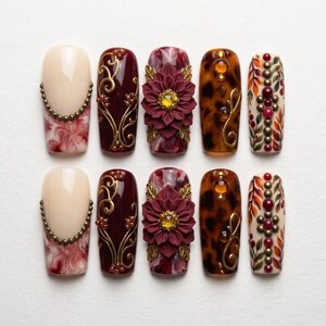 May include: A set of ten press-on nails featuring diverse designs including cream and red marble patterns with gold bead accents, deep burgundy with gold filigree, 3D burgundy flowers with yellow centers, brown tortoiseshell, and autumn leaf motifs.