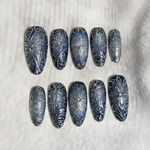 May include: A set of ten oval-shaped false nails. Each nail is painted with a dark blue base and intricate silver floral and swirl designs. The nails are arranged on a white surface.