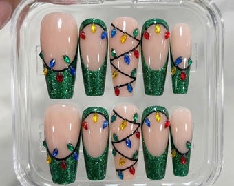 May include: A set of ten press-on nails in a clear plastic case. The nails feature a nude base with green glitter tips, black painted wire lines, and small colorful gemstone accents shaped like holiday light bulbs in red, blue, and yellow.
