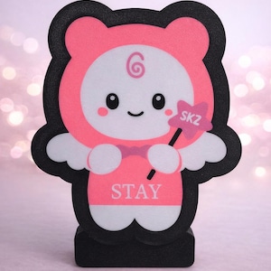 May include: A pink and black decorative figure in the shape of a cartoon character. The figure has a white face with a smiling expression, wearing a pink hooded outfit. It holds a star-shaped wand with "SKZ" on it, and the word "STAY" is printed on the chest.