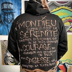 May include: Black hoodie with brown text on the back. The text reads "Mon Dieu, Donnez-moi la sérénité d'accepter les choses que je ne puis changer, le courage de changer les choses que je peux, et la sagesse d'en connaître la différence."