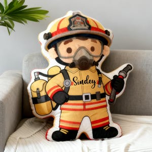 May include: A firefighter-themed decorative pillow in the shape of a cartoon character. The pillow features a yellow and red uniform, a helmet, and a breathing apparatus. The name "Sindey" is printed on the chest. The pillow is on a gray couch.