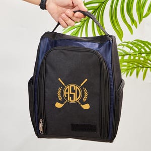 Puede incluir: Bolsa de zapatos de golf negra y azul marino con asa. La bolsa presenta un emblema dorado con palos de golf cruzados, una corona de laurel y las iniciales "ASD". La bolsa tiene bolsillos laterales y cierre de cremallera.