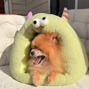Pode incluir: Um pequeno c&atilde;o Pomer&acirc;nia laranja boceja dentro de uma cama para animais de estima&ccedil;&atilde;o em forma de monstro verde claro. A cama tem olhos grandes e arregalados e chifres roxos e amarelos. A boca do c&atilde;o est&aacute; bem aberta, revelando sua l&iacute;ngua rosa.