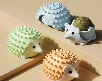 May include: Four colorful cat toys shaped like hedgehogs. The toys are in shades of light blue, green, orange, and white. One hedgehog holds a ball of dried catnip. The toys have a textured, spiky design.