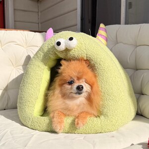 Pode incluir: Um pequeno c&atilde;o Pomer&acirc;nia laranja espreita de uma cama para animais de estima&ccedil;&atilde;o verde-claro em forma de monstro. A cama tem dois olhos grandes, orelhas cor-de-rosa e chifres listrados. A cama para c&atilde;es est&aacute; sobre uma almofada branca almofadada.