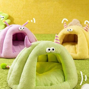 Pode incluir: Um close-up de tr&ecirc;s camas de estima&ccedil;&atilde;o de pel&uacute;cia em forma de monstros de desenho animado. As camas s&atilde;o em tons de verde, rosa e amarelo, com olhos grandes e arregalados e antenas em forma de chifre. As camas t&ecirc;m uma apar&ecirc;ncia macia e convidativa.