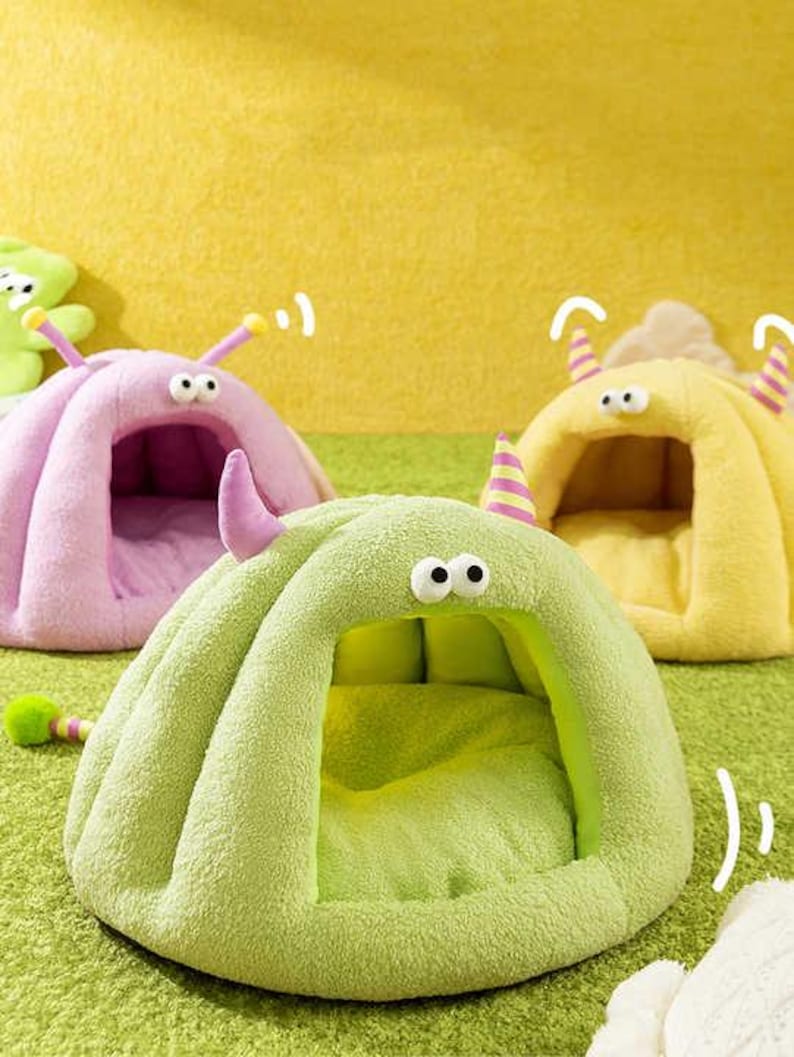 Pode incluir: Um close-up de tr&ecirc;s camas de estima&ccedil;&atilde;o de pel&uacute;cia em forma de monstros de desenho animado. As camas s&atilde;o em tons de verde, rosa e amarelo, com olhos grandes e arregalados e antenas em forma de chifre. As camas t&ecirc;m uma apar&ecirc;ncia macia e convidativa.