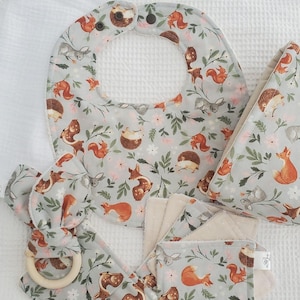 Peut inclure: Un ensemble d'accessoires pour bébé en tissu gris clair avec un motif d'animaux des bois incluant des renards, écureuils, hérissons et faons, agrémenté de petites fleurs roses et feuilles vertes. L'ensemble comprend un bavoir, un anneau de dentition.