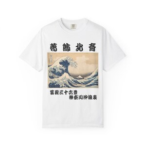 以下が含まれることがあります： 大きな波と山を描いた日本の木版画がプリントされた白いTシャツ。プリントは青、ベージュ、白を使用しています。日本語の文字が画像の上と下に印刷されています。