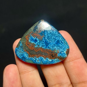 May include: A triangular polished gemstone featuring a vibrant mix of deep blue, turquoise, and earthy reddish-brown patterns. The surface displays a complex, swirling texture with metallic dark grey inclusions throughout the stone's face.