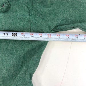 May include: Close-up of a green garment with a measuring tape laid across it. The tape shows measurements in inches, with markings from 11 to 20 inches. The fabric has a textured appearance.